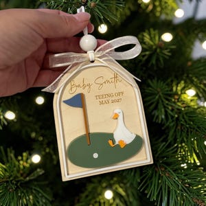 May include: An arched wooden ornament featuring a white goose on a green golf green with a blue flag and a white golf ball. The text reads Baby Smith TEEING OFF MAY 2027. The ornament is topped with a white bead and a sheer white ribbon for hanging.