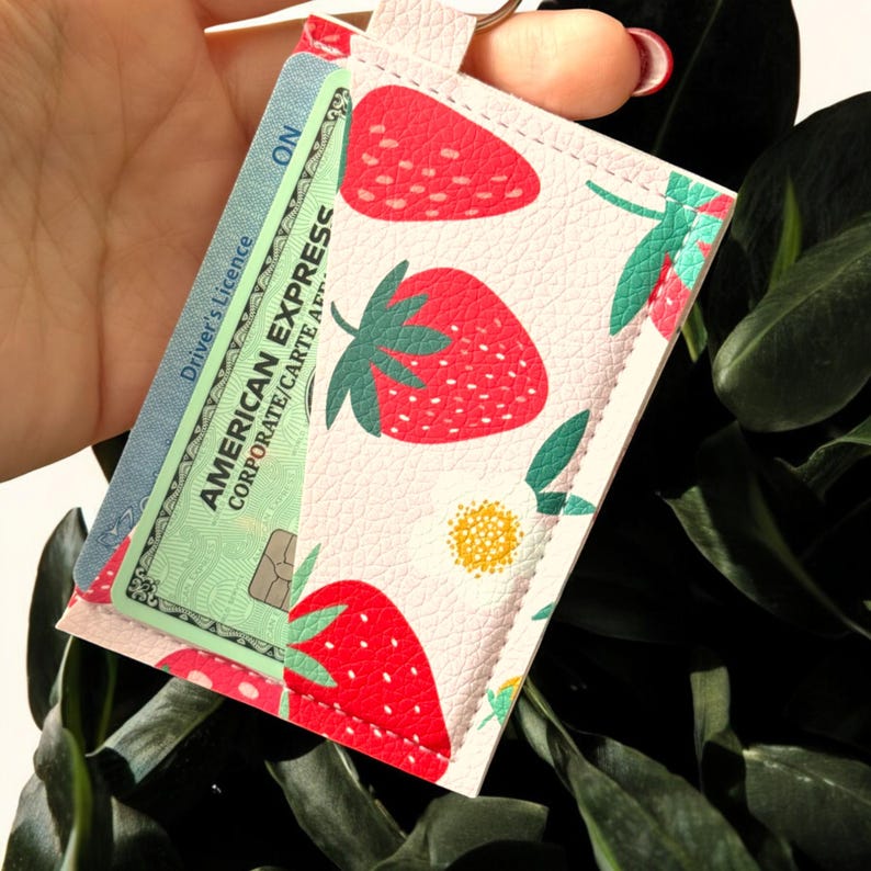 May include: A white leather card holder with a strawberry print. The card holder has a clear plastic window to display a credit card. The credit card is a green American Express card with the text "AMERICAN EXPRESS CORPORATE/CARTE APPR" and "Driver's Licence ON".