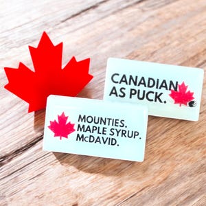 May include: Two white rectangular signs with black text and red maple leaf graphics. The top sign reads "Canadian as Puck." The bottom sign reads "Mounties. Maple Syrup. McDavid."
