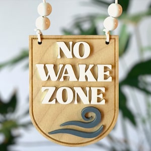 Peut inclure: Une pancarte en bois avec le texte "NO WAKE ZONE" en lettres blanches. La pancarte est suspendue à une chaîne de perles blanches et présente un motif de vague gris en bas.