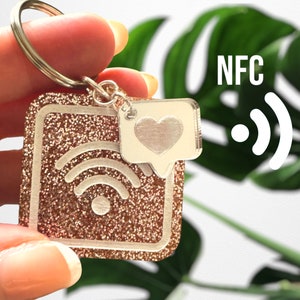 Peut inclure: Un porte-clés en paillettes or rose avec un symbole wifi blanc et un charme en forme de cœur argenté. Le porte-clés a un anneau argenté et le texte "NFC" est visible en arrière-plan.