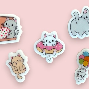 May include: Five colourful stickers featuring cute cartoon cats in various poses. One cat is holding a pizza slice, another is in a doughnut, one is flying with balloons, and two are sitting.