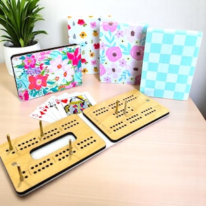 May include: A wooden cribbage board with a light wood finish and black edges. The board is open to reveal the full game in two laps. The board is on a light wood table with four playing cards and three decorative boxes with floral patterns.