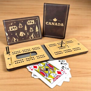 May include: A wooden cribbage board game set with a brown leather case featuring the word "CANADA" in gold lettering. The case also has a maple leaf design. The board is open and shows the pegs and scoring holes. There are also four playing cards on the table.