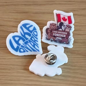 May include: Three white acrylic pins. One is a heart shape with blue text reading LAKE ONTARIO. Another features a beaver, a goose, and an otter holding a sign that says LAKE ONTARIO FOREVER under a Canadian flag. The third shows the metal pin back.