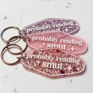 May include: Three motel-style keychains with metal rings. One is purple glitter, one is solid matte pink, and one is pink glitter. Each features the white text "probably reading" or "probably reading smut" with small star icons printed on the surface.