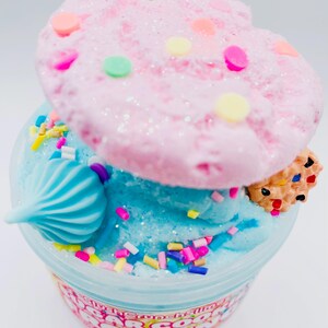 May include: A clear plastic container filled with blue slime, topped with a pink, glittery, cookie-shaped decoration and colorful sprinkles. A small, decorated cookie and a blue swirl are also on top. The container has text around the bottom.
