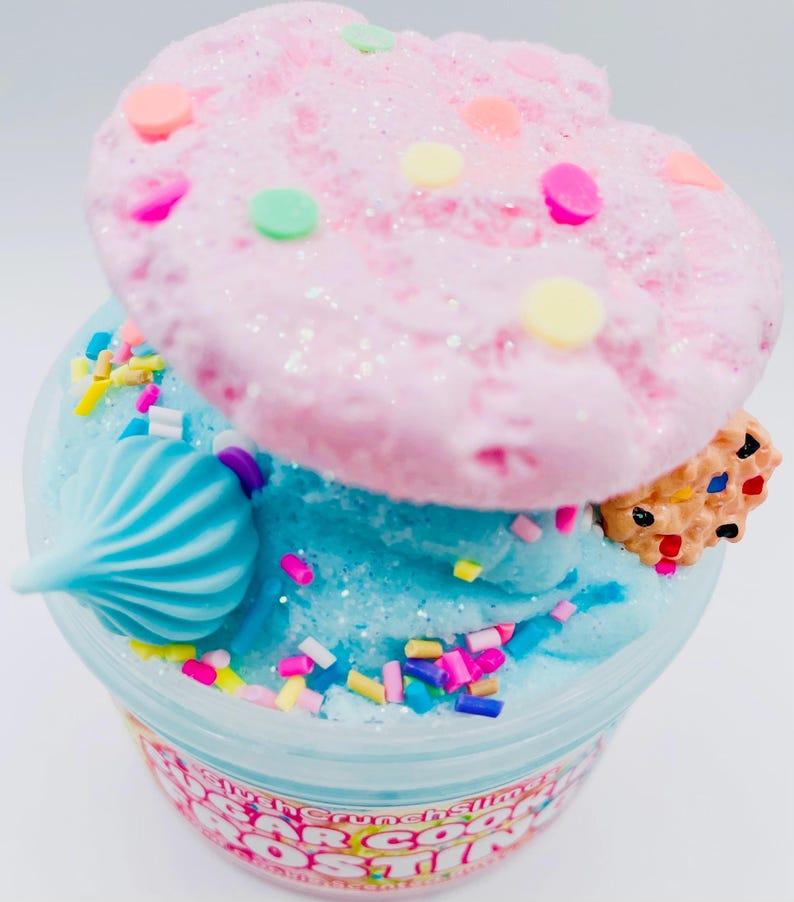 May include: A clear plastic container filled with blue slime, topped with a pink, glittery, cookie-shaped decoration and colorful sprinkles. A small, decorated cookie and a blue swirl are also on top. The container has text around the bottom.