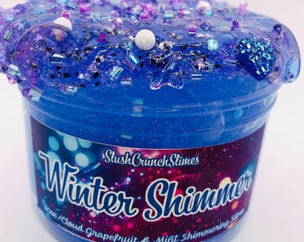 Op de afbeelding: Een doorzichtige plastic container gevuld met blauwe slijm, versierd met iriserende kralen, glitter en stervormige versieringen. Het etiket leest "Winter Shimmer" met het merk "SlushCrunch Slimes". De slijm heeft een glinsterend uiterlijk.