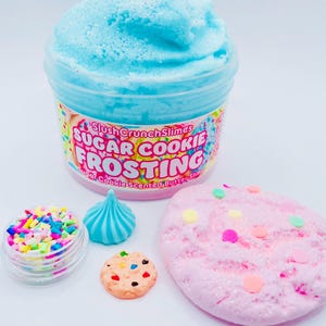 May include: A jar of blue, sparkly slime labeled "Sugar Cookie Frosting" with a variety of colorful sprinkles and miniature cookie-shaped decorations. A pink, glittery slime with colorful sprinkles is also present.