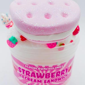 May include: A container of strawberry-scented cloud slime, resembling an ice cream sandwich. The top layer is pink with glitter and holes, the middle is white, and the base is clear with pink accents and strawberry-shaped decorations. The label reads "Strawberry Ice Cream Sandwich."