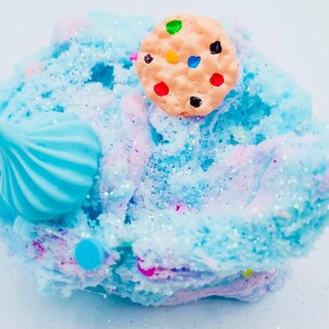 May include: A close-up of a colorful slime creation. The slime is a mix of blue, pink, and white, with glitter accents. A light brown cookie with colorful candy dots sits on top. A blue swirl and a blue dot are also visible.
