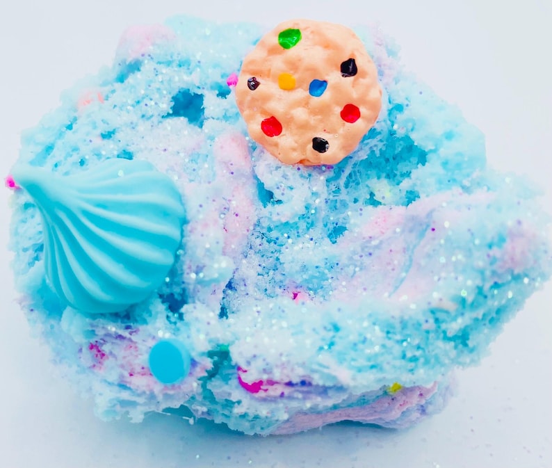 May include: A close-up of a colorful slime creation. The slime is a mix of blue, pink, and white, with glitter accents. A light brown cookie with colorful candy dots sits on top. A blue swirl and a blue dot are also visible.