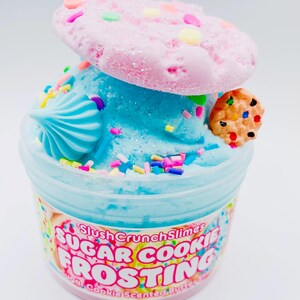 May include: A clear plastic container filled with blue slime, topped with a pink cookie and a small cookie. The container has text that reads "Sugar Cookie Frosting". Sprinkles and a blue swirl decorate the slime.