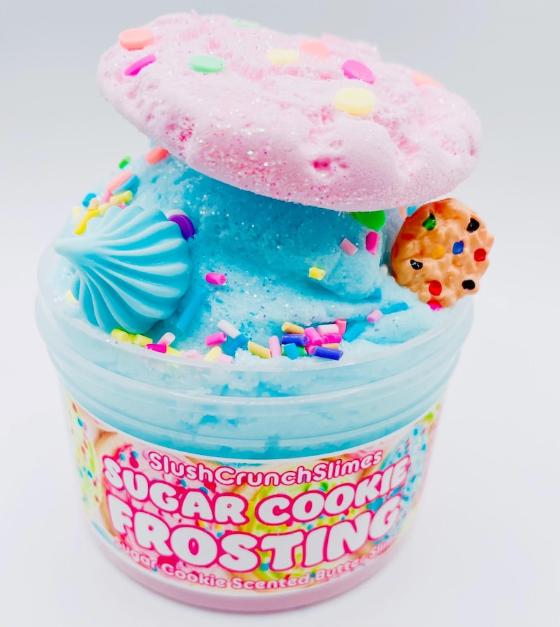 May include: A clear plastic container filled with blue slime, topped with a pink cookie and a small cookie. The container has text that reads "Sugar Cookie Frosting". Sprinkles and a blue swirl decorate the slime.
