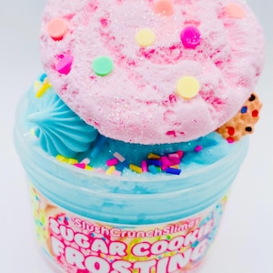 May include: A container of blue slime topped with a pink, glittery cookie-shaped decoration. The cookie has colorful circular sprinkles. The container has the text "Sugar Cookie Frosting".