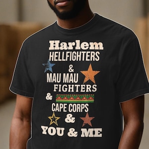 Può includere: Maglietta nera con una grafica in stile vintage che recita "Harlem Hellfighters & Mau Mau Fighters & Cape Corps & You & Me".
