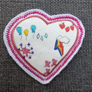 Può includere: Una toppa a forma di cuore con centro bianco e bordo cucito rosa. La superficie presenta illustrazioni disegnate a mano tra cui palloncini blu e gialli, piccoli diamanti blu, mele rosse, un fulmine arcobaleno e fiori rosa.
