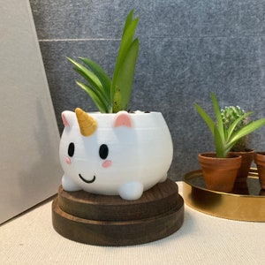 Peut inclure: Un pot en céramique blanc en forme de licorne avec une corne dorée et des oreilles roses. Le pot est rempli d'une plante succulente verte et repose sur un support en bois.
