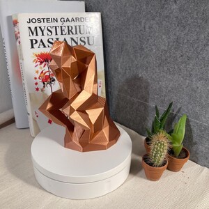 Puede incluir: Una escultura geométrica de color cobre de una persona en pose pensativa. La escultura está sobre un soporte giratorio blanco. Libros y pequeñas plantas en macetas están en el fondo.