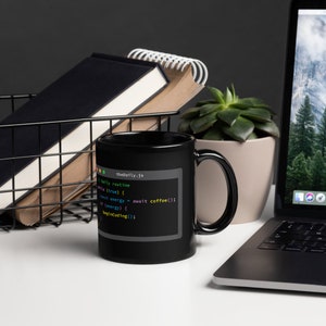K&ouml;nnte beinhalten: Schwarze Kaffeetasse mit einem wei&szlig;en Design eines Computerbildschirms, auf dem Code angezeigt wird. Der Code lautet "theDaily.js" und enth&auml;lt den Text "await coffee".