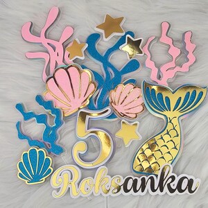 Op de afbeelding: Een caketopper met een zeemeermin-thema met een gouden nummer 5, een zeemeerminstaart, schelpen en zeewier. De topper bevat ook de naam "Roksanka" in gouden letters.