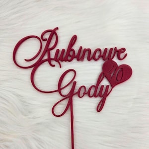 Può includere: Un cake topper bordeaux con le parole "Rubinowe Gody" in corsivo. Una forma di cuore con il numero "40" è sul lato destro. Il topper è su una superficie bianca strutturata.