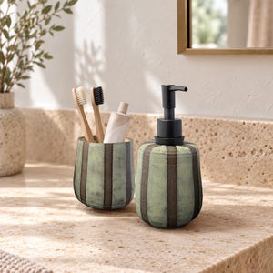 May include: A ceramic bathroom set featuring a soap dispenser with a black pump and a matching tumbler. Both items have a textured, sage green and dark brown vertical striped pattern. The tumbler holds two bamboo toothbrushes and a tube of toothpaste.