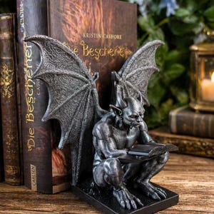 May include: A detailed, dark gray gargoyle bookend, with large wings, is reading a book. The bookend is on a black base, and is positioned next to a stack of books. The books have gold lettering.