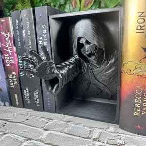 May include: A black, 3D-printed bookend featuring a skeletal figure reaching out from a rectangular frame. The figure has a hooded cloak and detailed facial features. Books are visible on either side of the bookend, creating a unique display.
