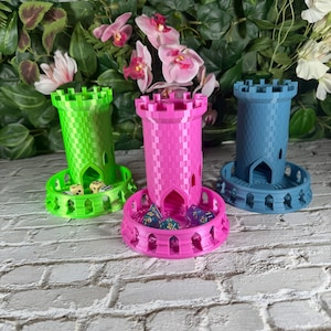 Könnte beinhalten: Drei 3D-gedruckte, turmförmige Würfeltabletts, eines grün, eines rosa und eines blau. Jeder Turm hat eine kleine Öffnung unten, damit die Würfel herausrollen können. Die Türme stehen auf einer weißen Ziegelstein-Oberfläche.