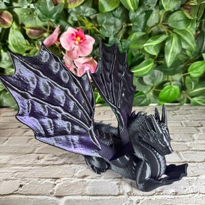 May include: A black dragon figurine with detailed wings featuring purple accents. The dragon is posed on a white brick surface, with a backdrop of green foliage and pink flowers. The figurine is a fantasy-themed decorative item.