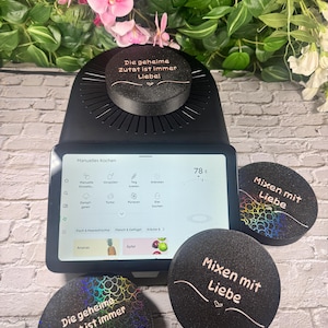 May include: A black kitchen appliance with a digital display, topped with a round lid and surrounded by three matching coasters. The coasters and lid feature the text "Die geheime Zutat ist immer Liebe!" and "Mixen mit Liebe".