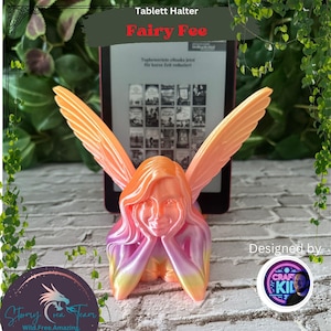 May include: A 3D printed fairy figurine with pink, orange, and yellow wings, holding her head in her hands. The fairy is sitting on a table next to a tablet stand. The text "Tablett Halter Fairy Fee" is above the fairy. The text "Designed by CRAFT KID" is below the fairy.