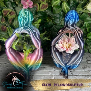 以下が含まれることがあります： 花瓶を持った女性の虹色の青と紫の樹脂彫刻が2つあります。彫刻は座った姿勢で、手は花瓶の周りを抱きしめています。彫刻には「エルフィン プランツェンスカルプチュール」とラベルが貼られています。