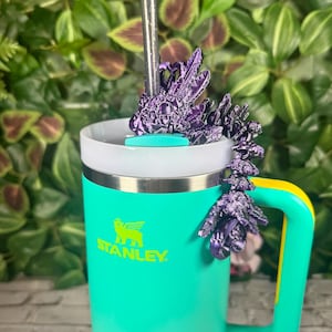 Pode incluir: Um copo Stanley turquesa com aro prateado e alça amarela. Uma decoração roxa em forma de cristal adorna a tampa, com um canudo prateado. O logotipo da Stanley é impresso em verde. O fundo é um verde exuberante.