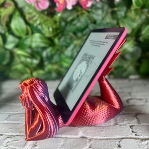 May include: A red mermaid-shaped phone or tablet stand. The stand is holding a tablet with a black and white screen. The stand is on a white brick surface.