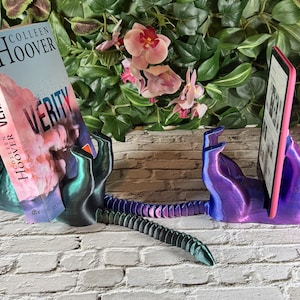 May include: A pair of cat-shaped book and tablet holders. One holds a book titled "Verity" by Colleen Hoover. The other holds a tablet. The holders are iridescent, with a gradient of green, blue, and purple.