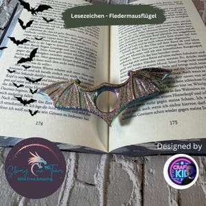 May include: A colorful iridescent bat-shaped bookmark with glitter accents. The bookmark is designed to be used as a finger ring. The bookmark is open on a book with the text "Lesezeichen - Fledermausflügel" visible.