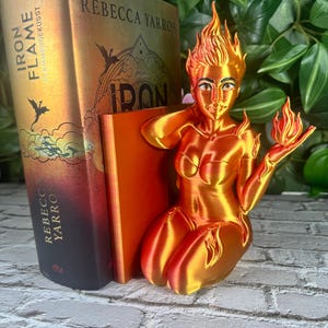 May include: A 3D-printed orange and yellow figurine of a woman with flames for hair and holding a flame. The figurine is next to a book with the title "Iron Flame" and the author's name, Rebecca Yarros.