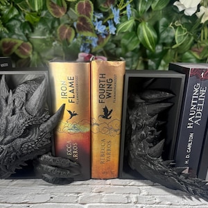 Könnte beinhalten: Schwarze Buchstützen im Drachen-Design halten Bücher mit Titeln wie "Iron Flame" und "Fourth Wing". Die Buchstützen sind mit Drachenköpfen und -krallen detailliert. Die Bücher haben goldene und rote Einbände.