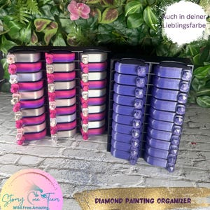 Könnte beinhalten: Zwei Diamant-Mal-Organizer, einer mit rosa und weißen Fächern und der andere mit violetten Fächern. Die Organizer sind aus schwarzem Kunststoff und haben ein rautenförmiges Design auf der Vorderseite. Der Text "DIAMANT-MAL-ORGANIZER" ist unter den Organisatoren gedruckt.
