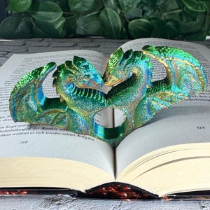 May include: A heart-shaped book page holder featuring two dragons in shades of green, blue, and gold. The dragons are positioned to embrace, with their heads touching. The holder is resting on an open book, with the page numbers 318 and 319 visible.