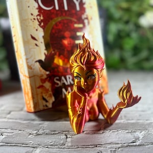 May include: A 3D-printed mermaid bookend in shades of gold and red, resting on a white brick surface. The mermaid is posed with her chin on her hand, with a book titled "CRESCENT CITY" in the background.