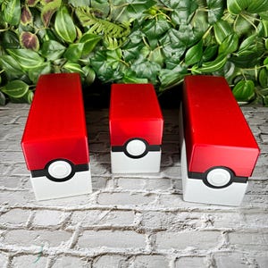 Könnte beinhalten: Drei rote und weiße Pokémon-Sammelkartenboxen mit einem Pokéball-Design. Die Boxen sind in verschiedenen Größen und in einer Reihe auf einer weißen Ziegelstein-Oberfläche angeordnet.