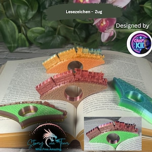 May include: Colorful train-shaped bookmarks in orange, red, and green, resting on the pages of a book. The bookmarks have a circular opening. The text "Lesezeichen - Zug" is visible.