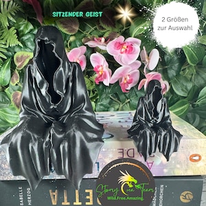 May include: Two black bookends shaped like hooded figures, sitting on a book. The larger bookend is on the left and the smaller bookend is on the right. The text "SITZENDER GEIST" is visible above the larger bookend. The text "2 Größen zur Auswahl" is visible above the smaller bookend.