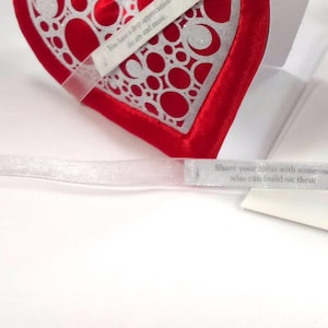 May include: A red velvet heart-shaped card with a white lace design. The card is open to reveal a white interior with a smaller heart-shaped cutout. The card is decorated with a white ribbon.
