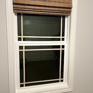 May include: A white-framed window with a woven bamboo blind at the top. The window features a grid pattern made with light-coloured tape. The room's interior is dark, with a glimpse of a door.