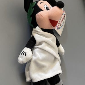 May include: A plush Mickey Mouse toy wearing a white toga and a green laurel wreath. The toy has a red mouth and black eyes. The tag on the toy reads "Disney Store".